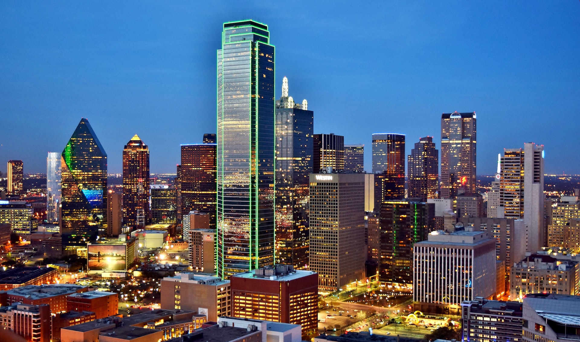 A tall glass skyscraper glows green in downtown Dallas